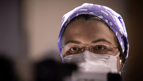 a close-up shot of Emanuela Binello as she performs surgery. She is wearing a royal purple scrub cap with white dots, glasses and a white mask.